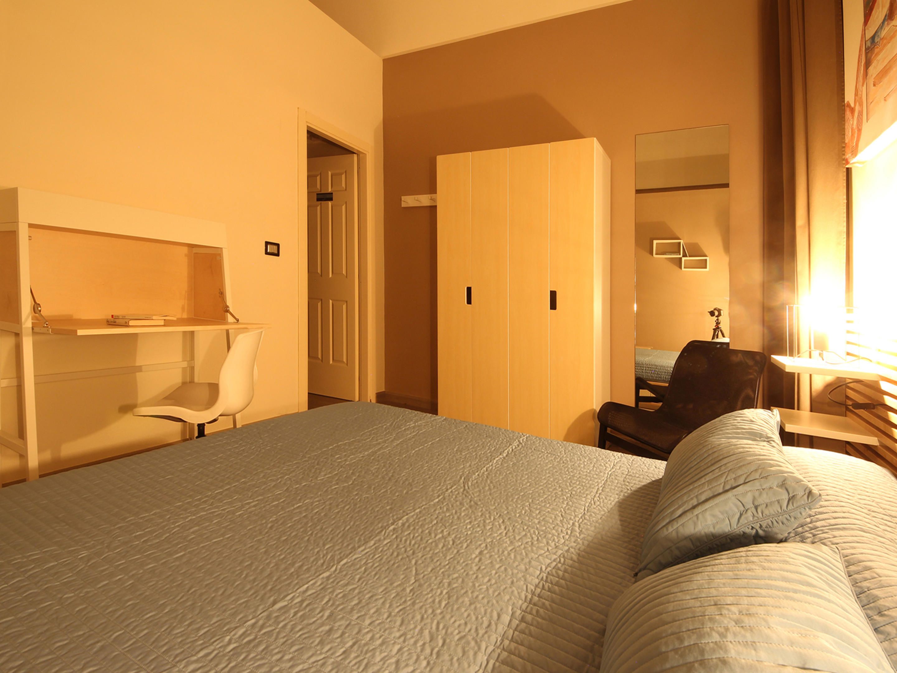 A bedroom with a bed, a desk, a chair, a wardrobe, and a mirror.