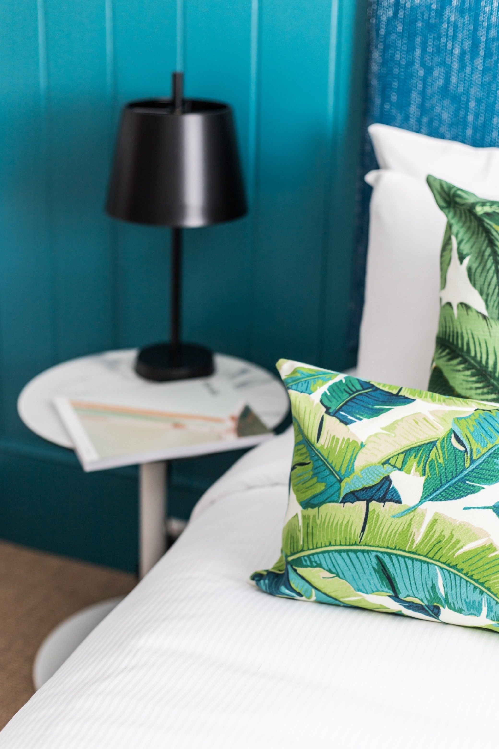 A neatly made bed with a white pillow and a green leaf-patterned pillow. A small white table with a black lamp and a magazine is beside the bed. The room has teal walls.