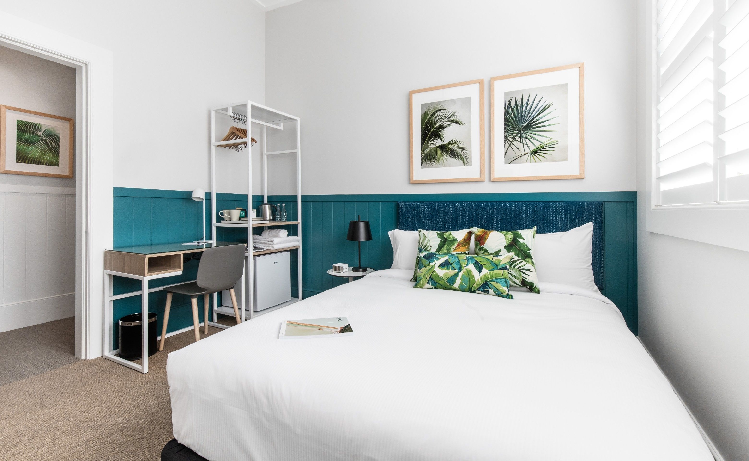 A hotel room with a bed, a desk, a chair, a wardrobe, and two framed pictures on the wall.