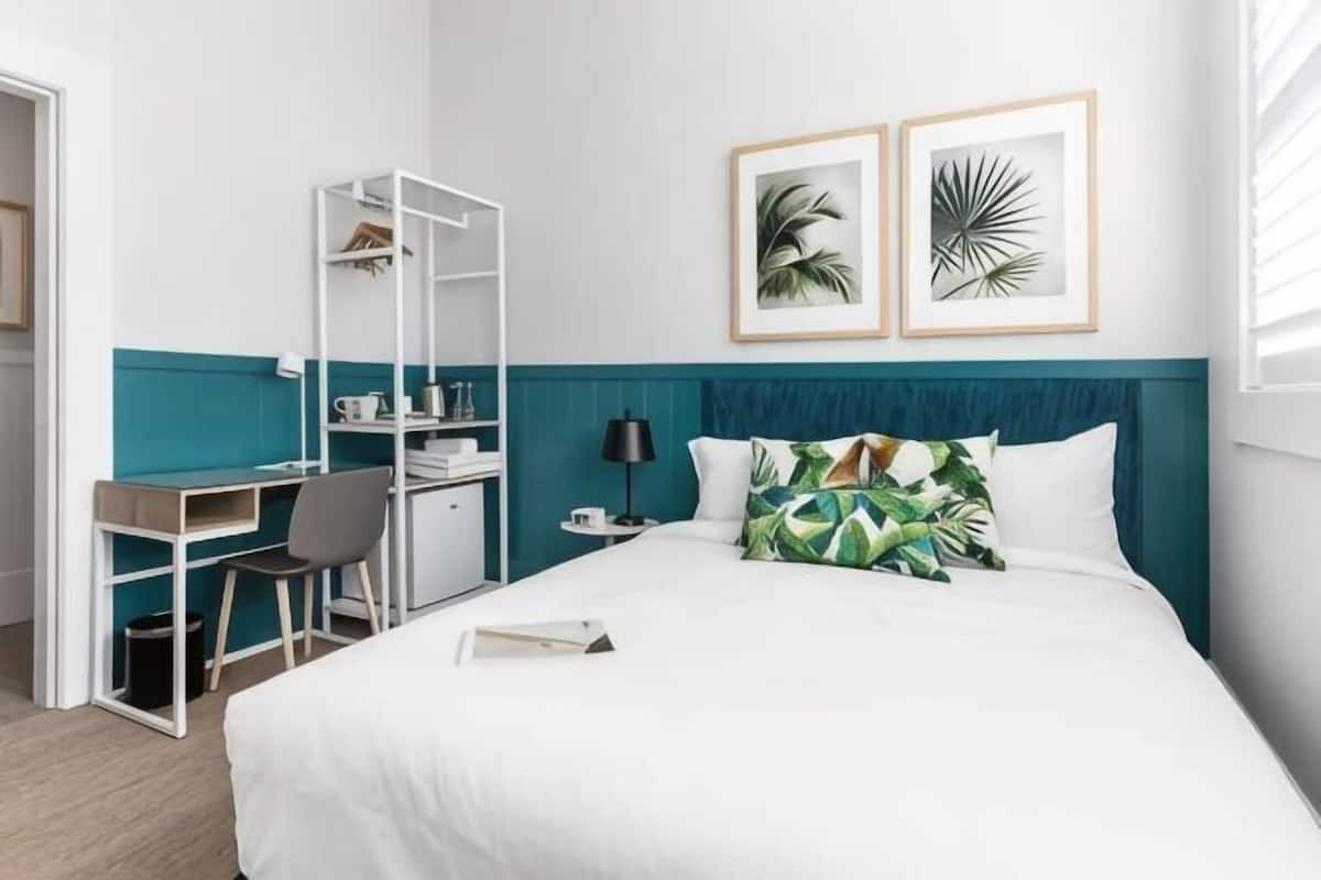 A hotel room with a bed, a desk, a chair, and a bookshelf.