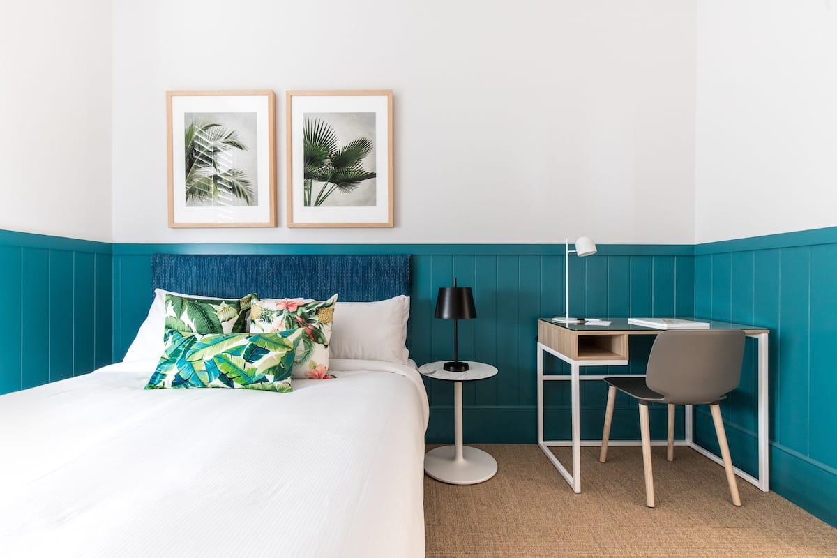 A hotel room with a bed, a desk, and two framed pictures on the wall.