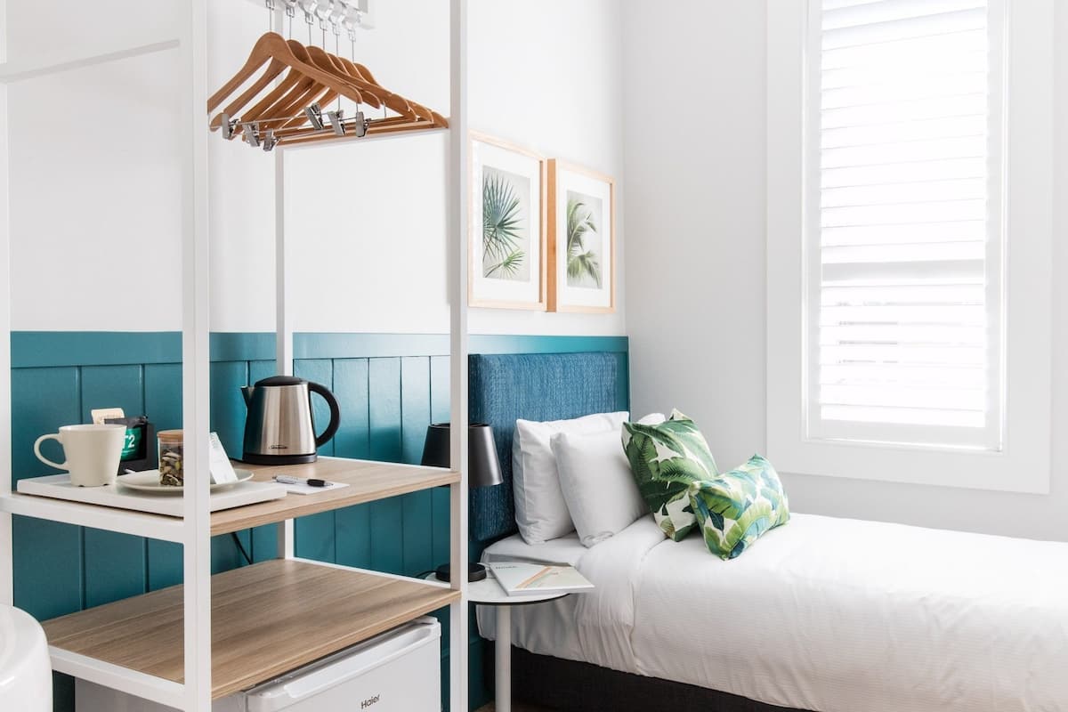 A compact hotel room with a bed, a small kitchenette, a white wardrobe, and a window with blinds.