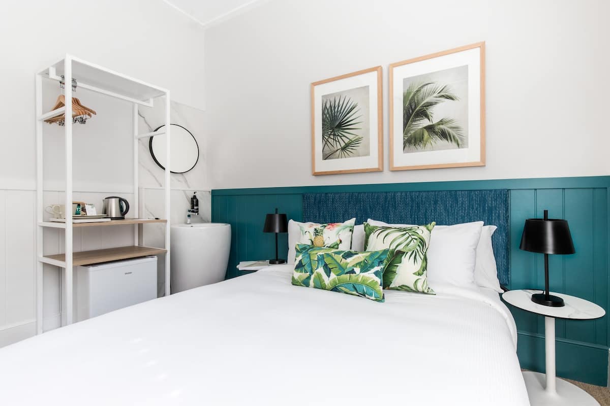 A hotel room with a bed, a wardrobe, a small table, a lamp, and two framed pictures on the wall.