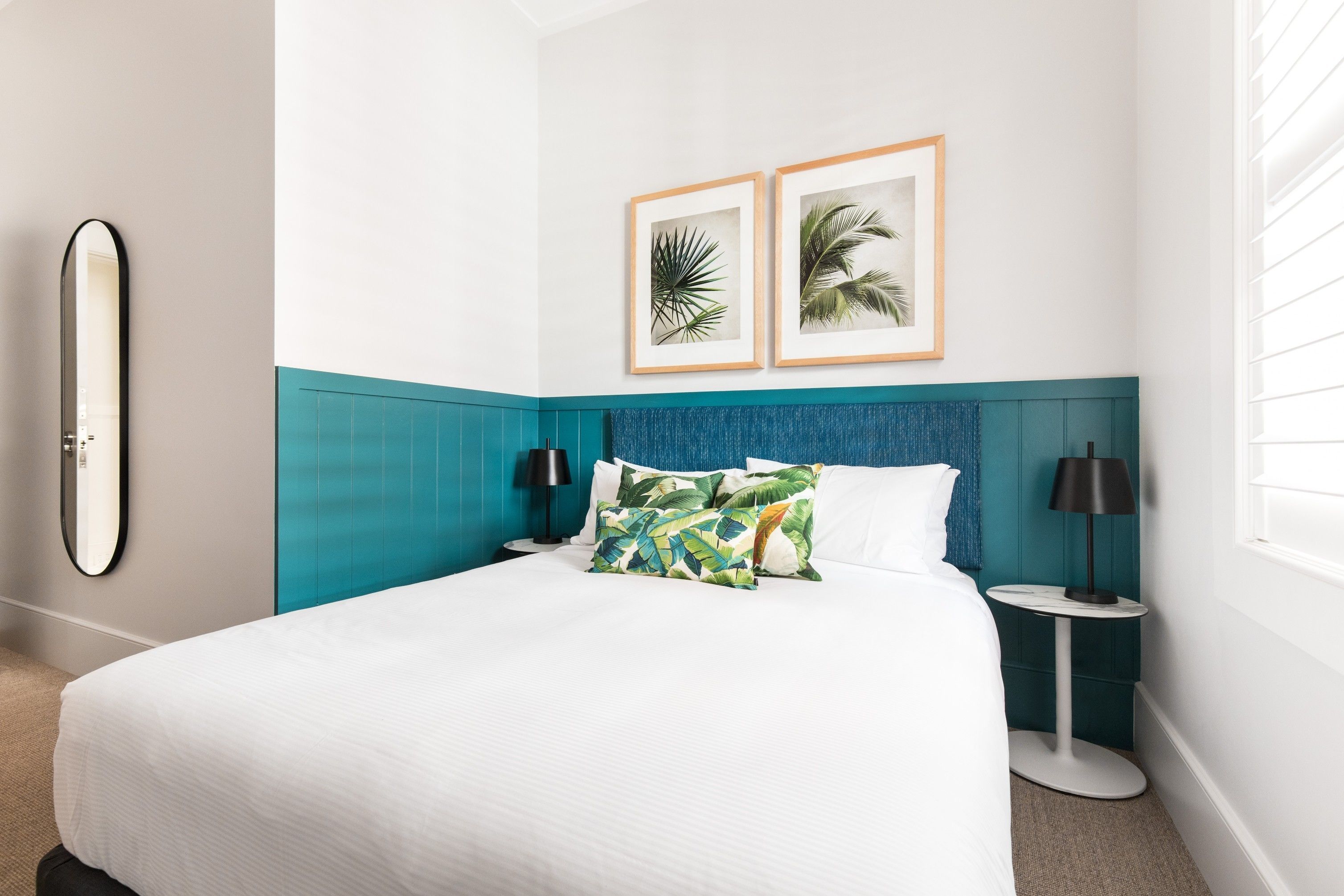 A neatly made bed with white linens, a teal headboard, and tropical-themed pillows. There are two framed pictures on the wall, a round mirror, and a small bedside table with a lamp.
