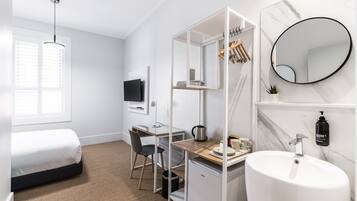 Quarto de hotel compacto com cama, escrivaninha com cadeira, pequeno espaço de cozinha, banheiro com pia e espelho, e janela com persianas.