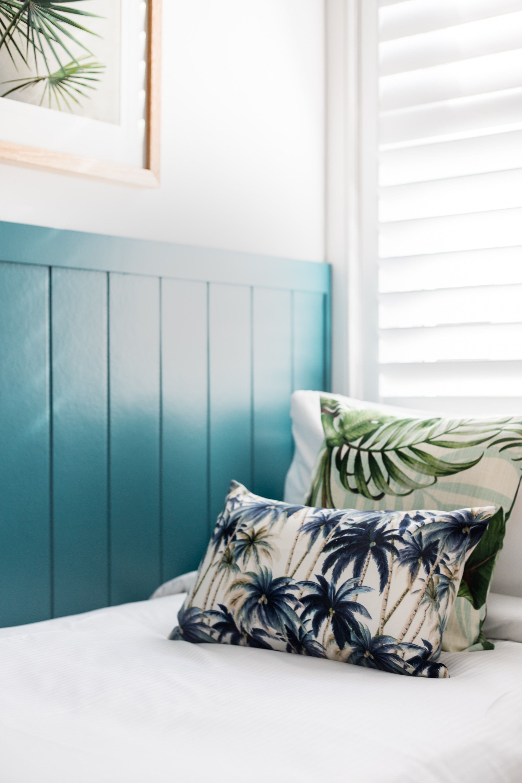 A bed with a teal headboard, a pillow with a tropical leaf pattern, and a framed picture on the wall.