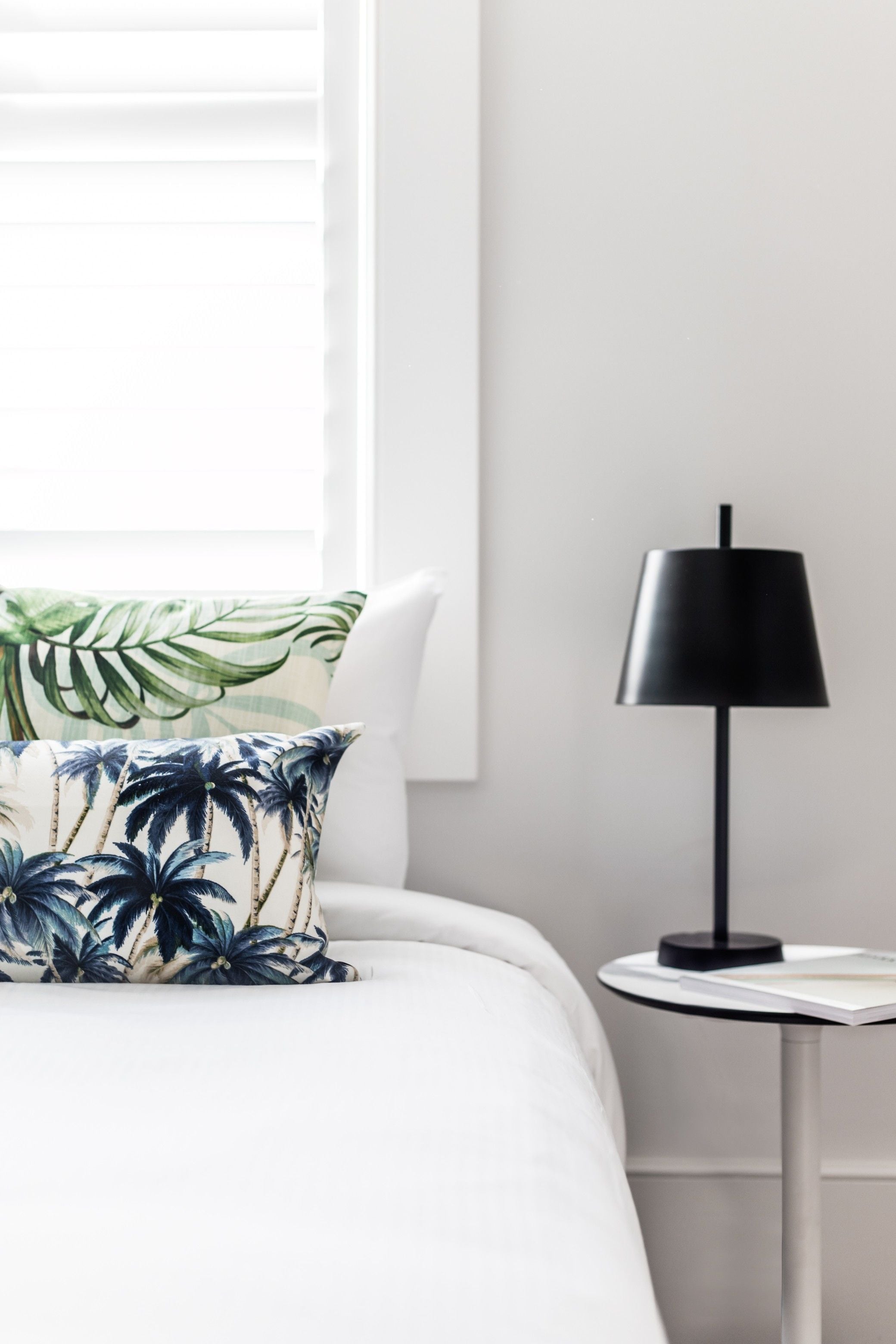 Una cama bien hecha con una colcha blanca y un cojín con motivos de hojas tropicales. Una lámpara de pie negra está sobre una mesita blanca junto a la cama.