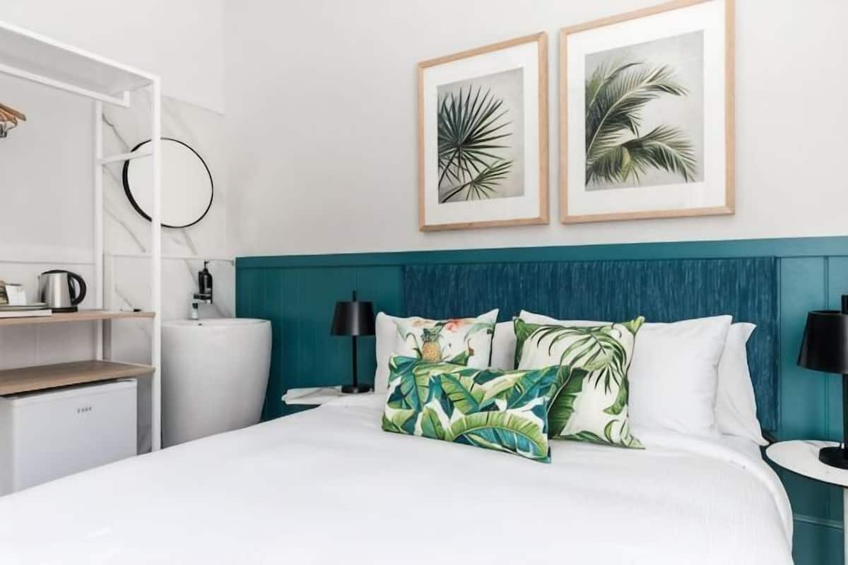 A hotel room with a bed, two framed plant artworks, a teal headboard, and a bedside table with a lamp.