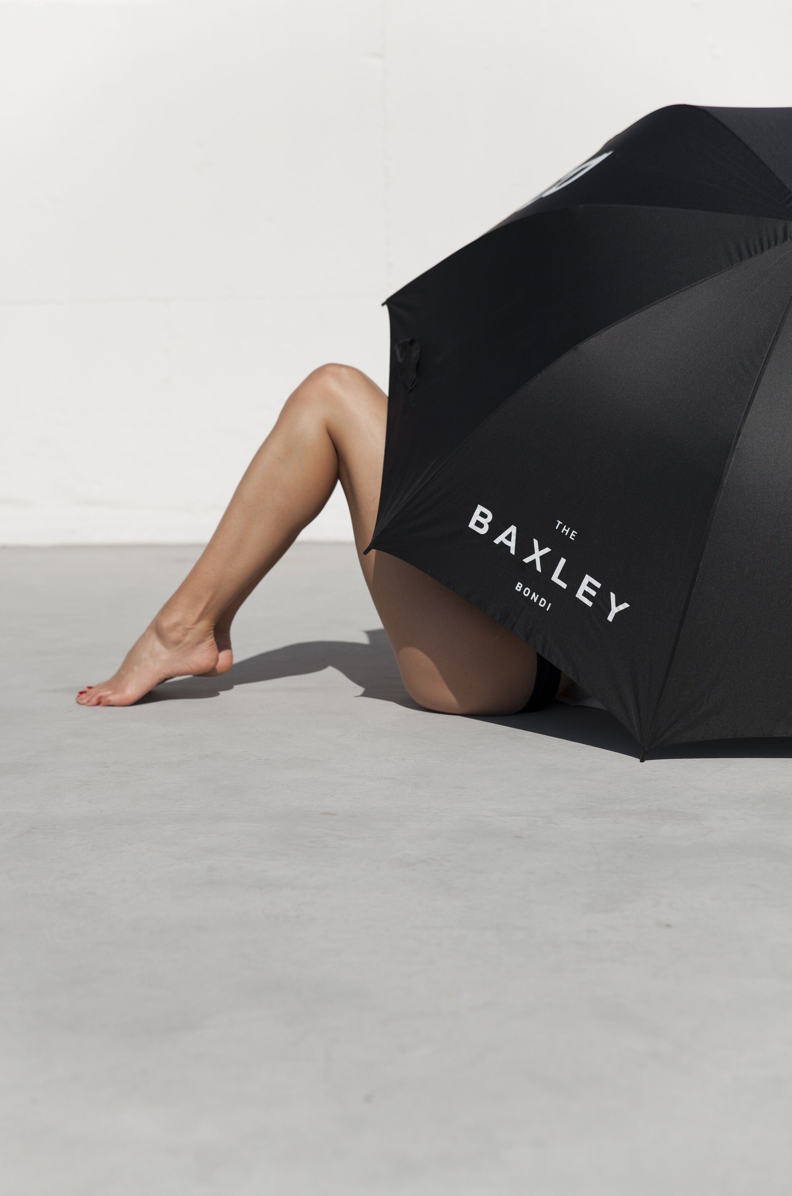 A person sitting with legs crossed, partially covered by a black umbrella with the text "THE BAXLEY BOND" on it.