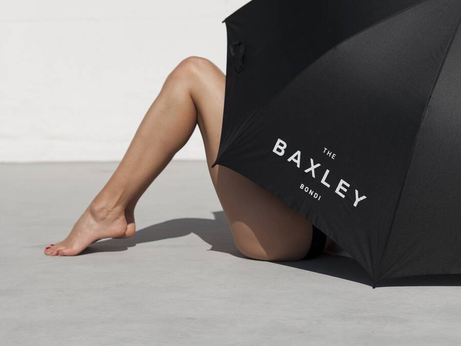 A person sitting with legs crossed, partially covered by a black umbrella with the text "THE BAXLEY BOND" on it.