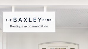 A white exterior of The BAXLEY BONDI Boutique Accommodation with a wooden floor, a white door with glass panels, and a wicker chair with a potted plant. - The Baxley Bondi (Bondi)