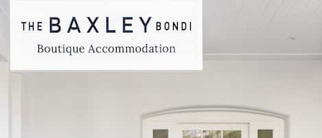 A white exterior of The BAXLEY BONDI Boutique Accommodation with a wooden floor, a white door with glass panels, and a wicker chair with a potted plant.