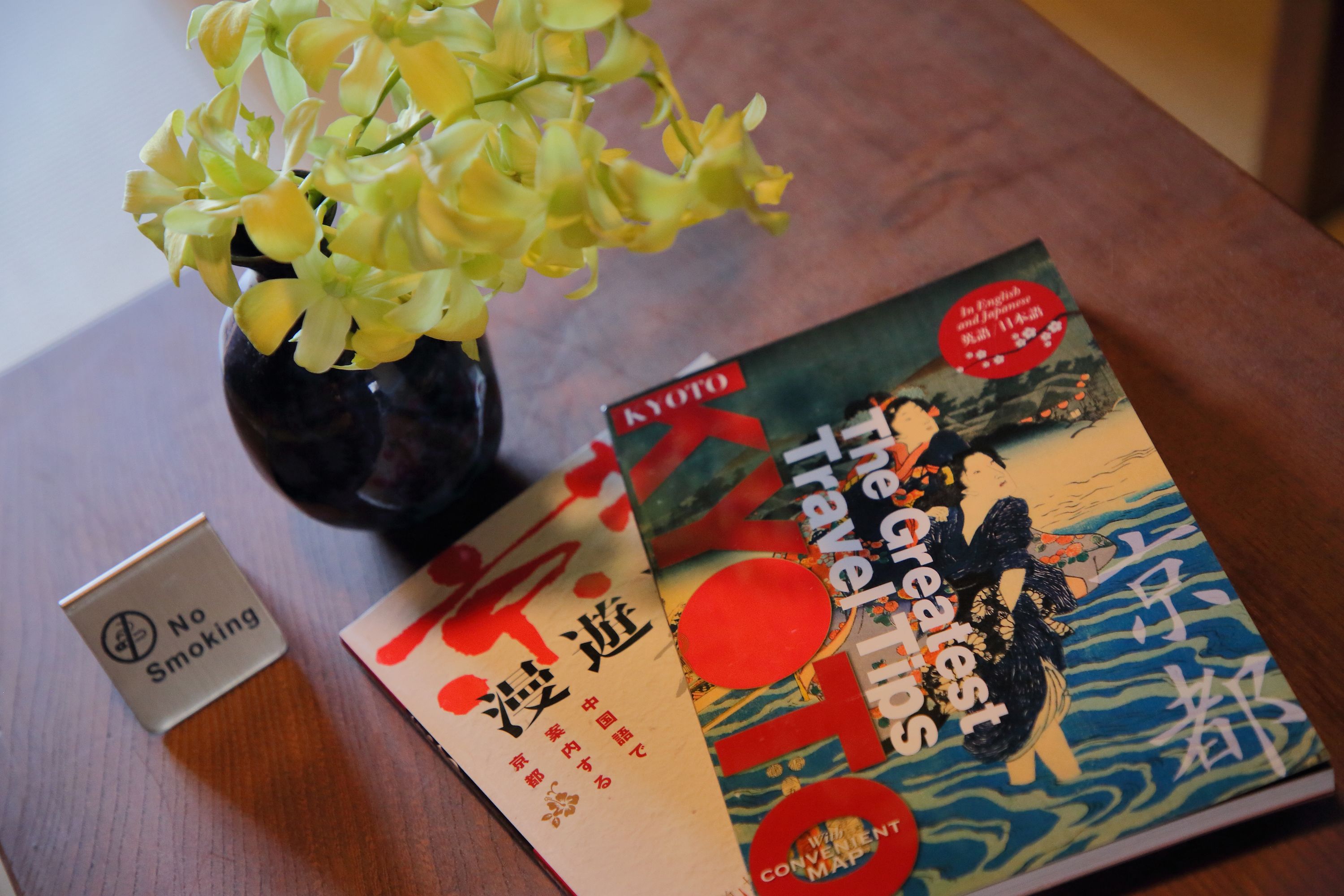 Een tafel met een vaas gele bloemen, een ‘Niet Roken’ bord en een boek getiteld 'The Greatest Travel Times'.