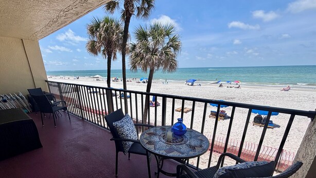 A balcony with a table and chairs, overlooking a beach with palm trees and umbrellas. - Direct Gulf Front Las Brisas 105, Very Nicely Updated 3 Bedroom/2 Bath (Madeira Beach)