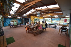 Un espacio comedor al aire libre cubierto, con mesas y bancos de madera, un mural en la pared y vistas al exterior.