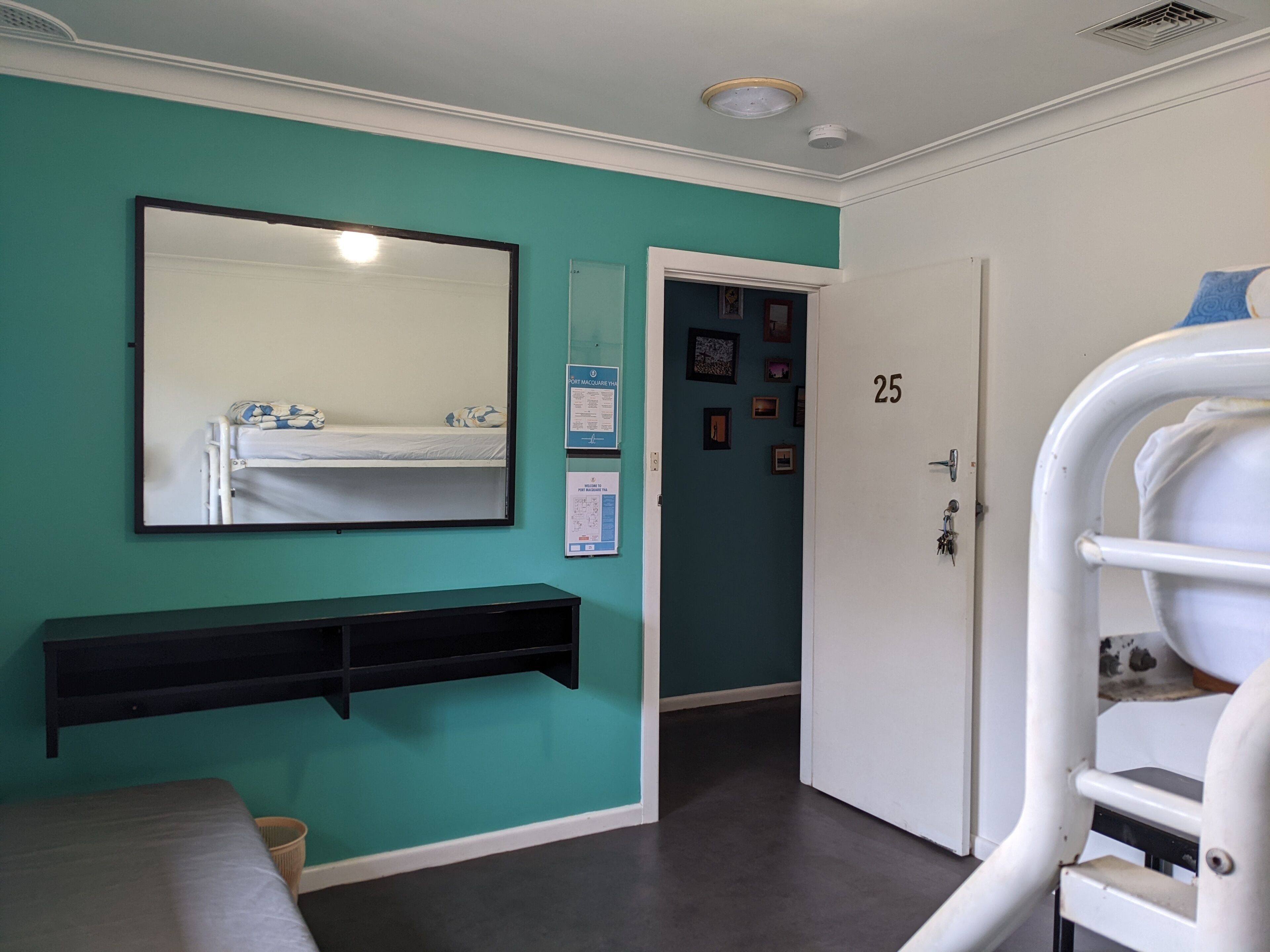 A room with teal walls, a white door marked with the number 25, a bed with a white mattress, a black bedside table, and a mirror on the wall.