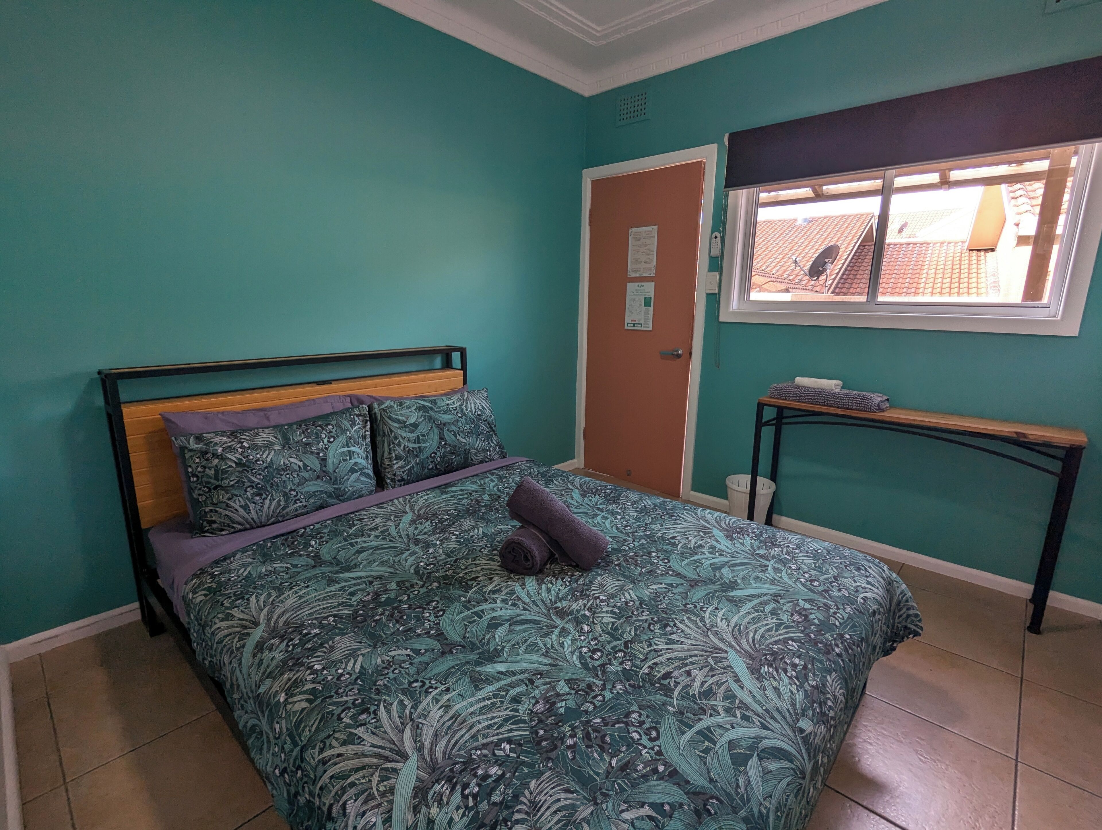 A bedroom with teal walls, a bed with a floral bedspread, a wooden headboard, and a window with a view of a tiled roof.