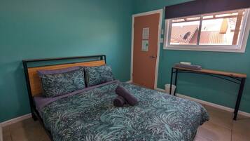 A bedroom with teal walls, a bed with a floral bedspread, a wooden headboard, and a window with a view of a tiled roof.