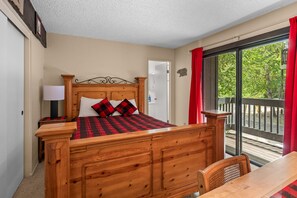A bedroom with a wooden bed, a nightstand, a lamp, and a view of the outdoors through a sliding glass door. - Lakeside Retreat Condo - Walk to Lake, Fireplace, Deck, Swimming Pool (Big Bear Lake)