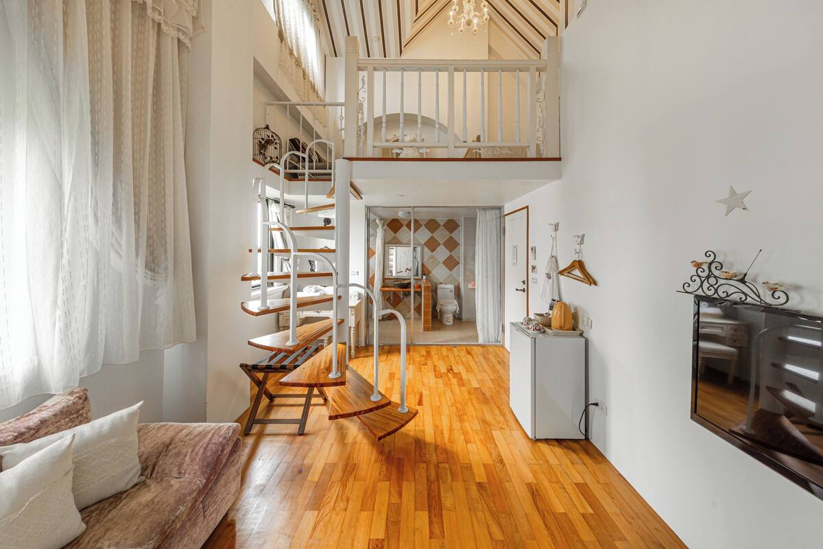Una habitación amplia con piso de madera, un sofá, un sillón mecedor, una escalera y una pequeña kitchenette.