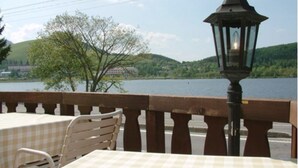 A table with a checkered tablecloth, a chair, and a lantern on a balcony overlooking a lake.