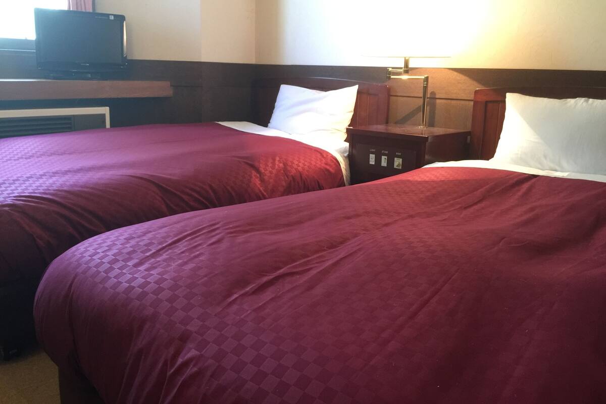 A hotel room with two beds, each with a red bedspread, a wooden headboard, and a bedside table with a lamp.