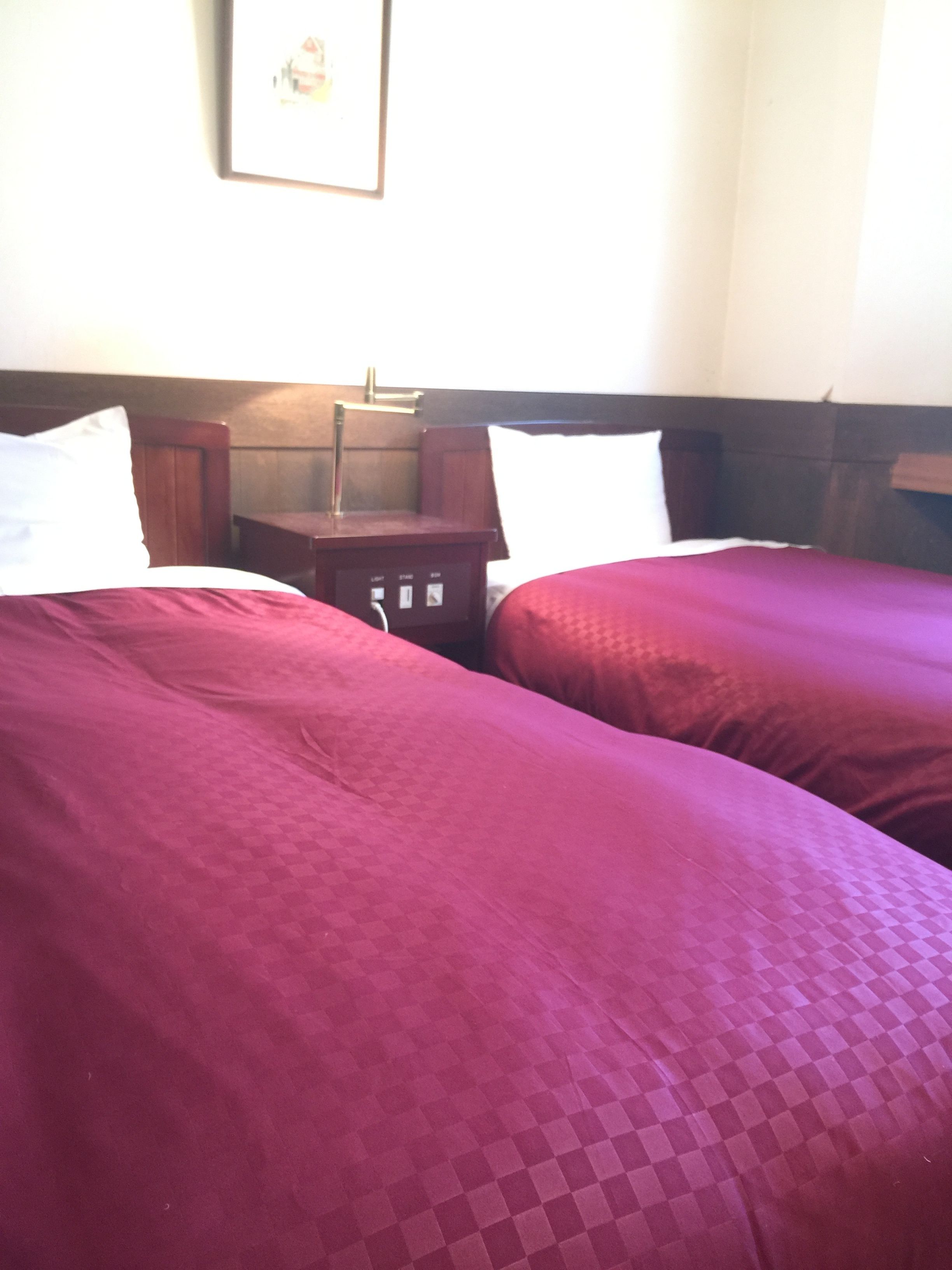 Habitación de hotel con dos camas, cada una con colcha roja y almohadas blancas. Entre las camas, una mesita de noche de madera con una lámpara.