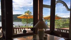 A table with a lamp and decorative items, a window with a view of a lake and outdoor seating with umbrellas.