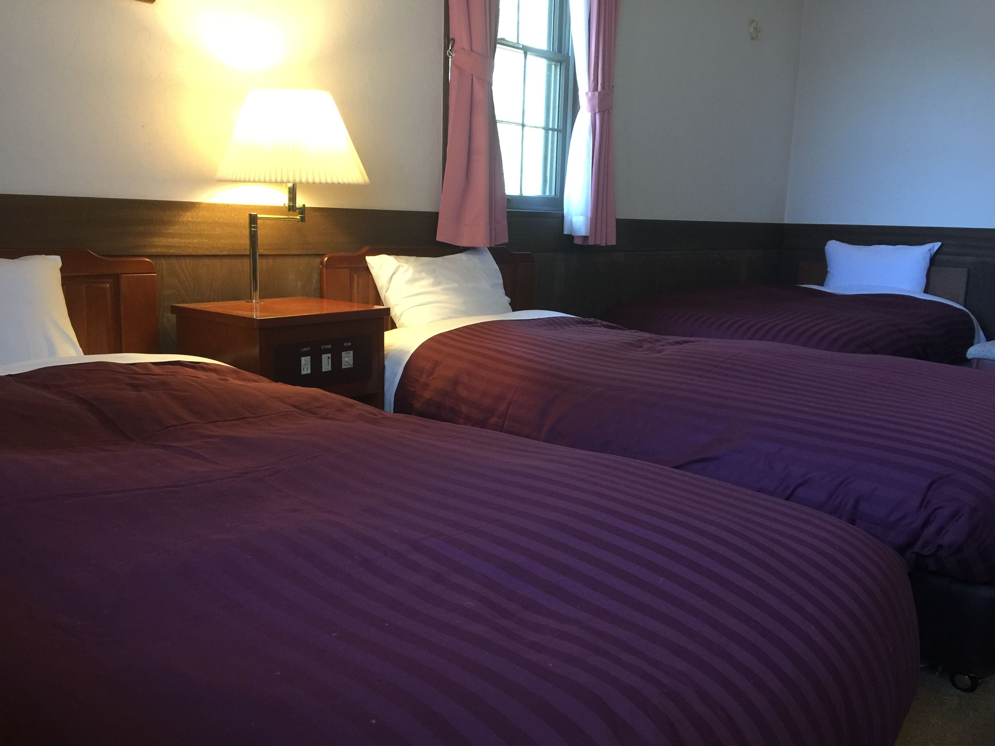 A hotel room with two beds, a bedside table, a lamp, and a window with curtains.