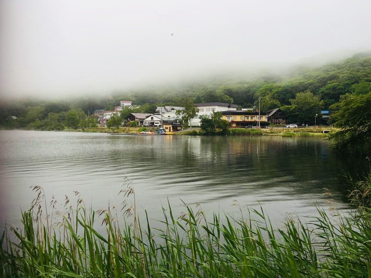 A calm lake with buildings on the shore, surrounded by greenery and fog.