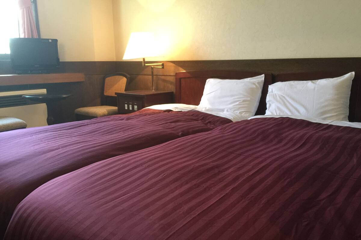 A hotel room with two beds, a desk, a chair, and a lamp.