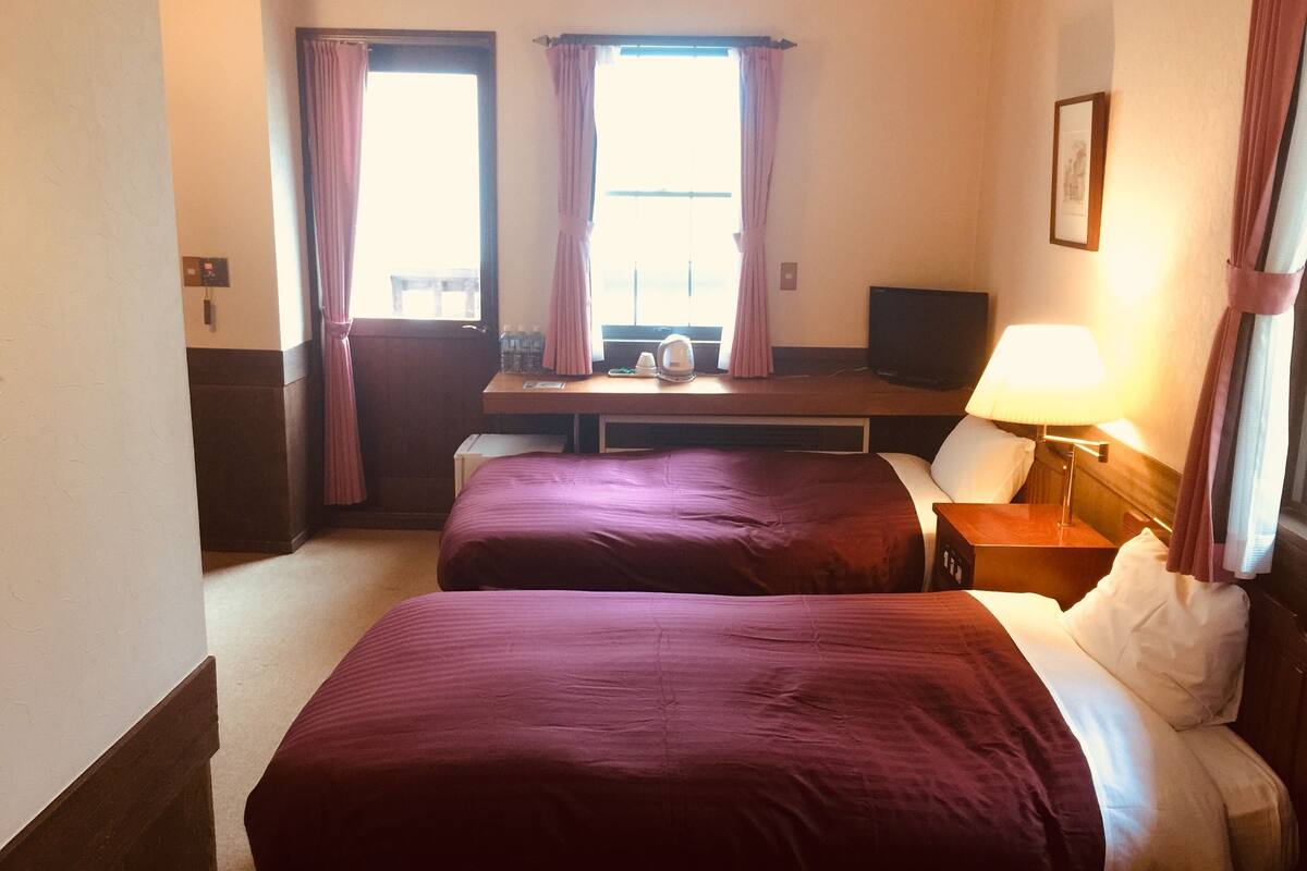 A hotel room with two beds, a desk, a TV, and a window with curtains.