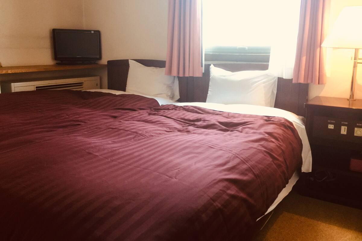A hotel room with a large bed, a television on a stand, a desk with a lamp, and a window with curtains.