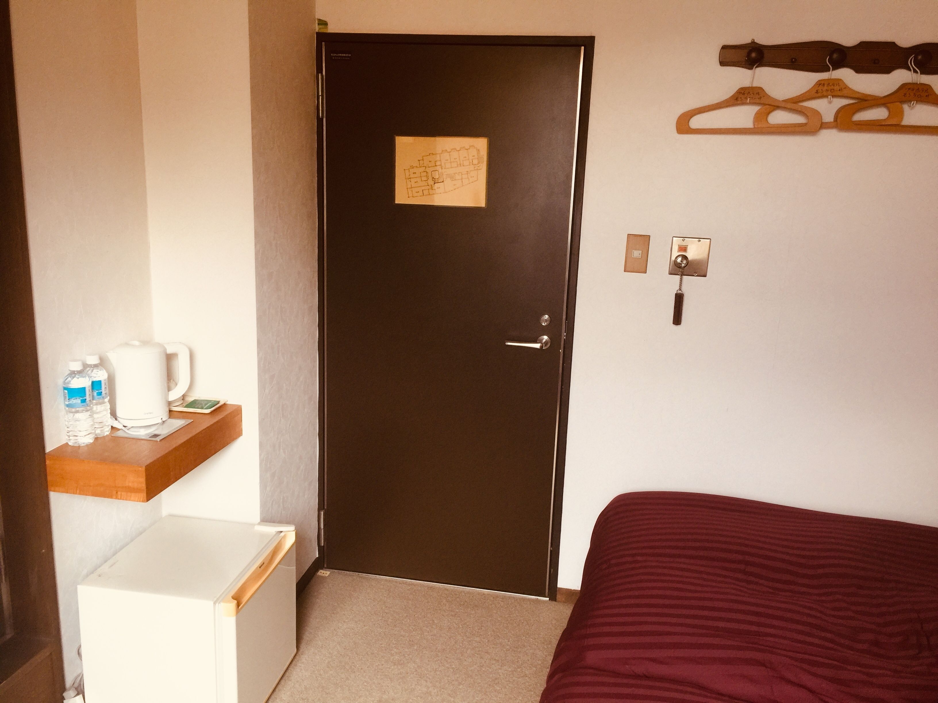 A hotel room with a dark door, a small white fridge, a wooden shelf with a kettle and water bottles, a bed with a red cover, and a coat rack with hangers.