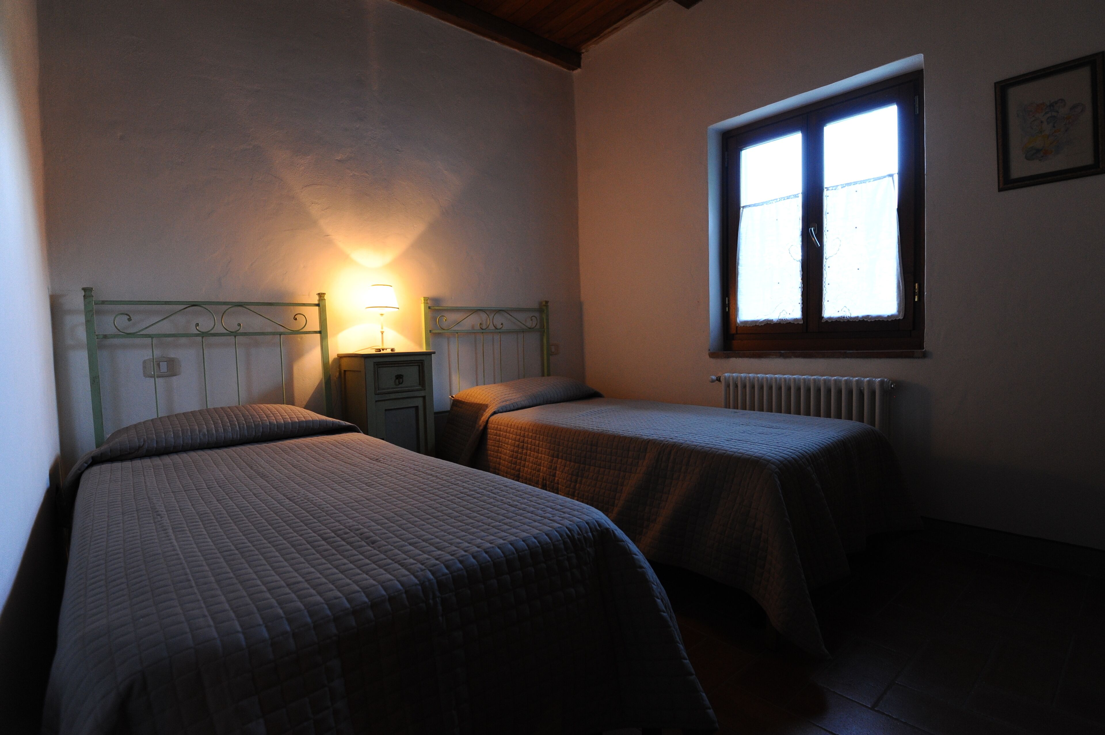 Un dormitorio con dos camas individuales, una mesita de noche con lámpara, una ventana con persianas y un radiador.
