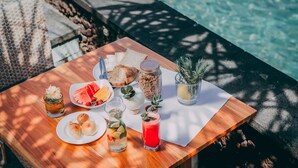 A wooden table with food and drinks by a poolside, including bread, fruit, and a drink in a glass. - Alam Candi Resort (Karangasem)