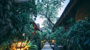 A narrow pathway flanked by lush greenery and traditional architecture, leading to a building with a thatched roof. - Alam Candi Resort (Karangasem)