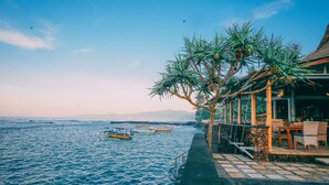 Ein Restaurant am Wasser mit Außenbestuhlung mit Blick auf das Meer, im Hintergrund Schiffe und ein klarer Himmel.