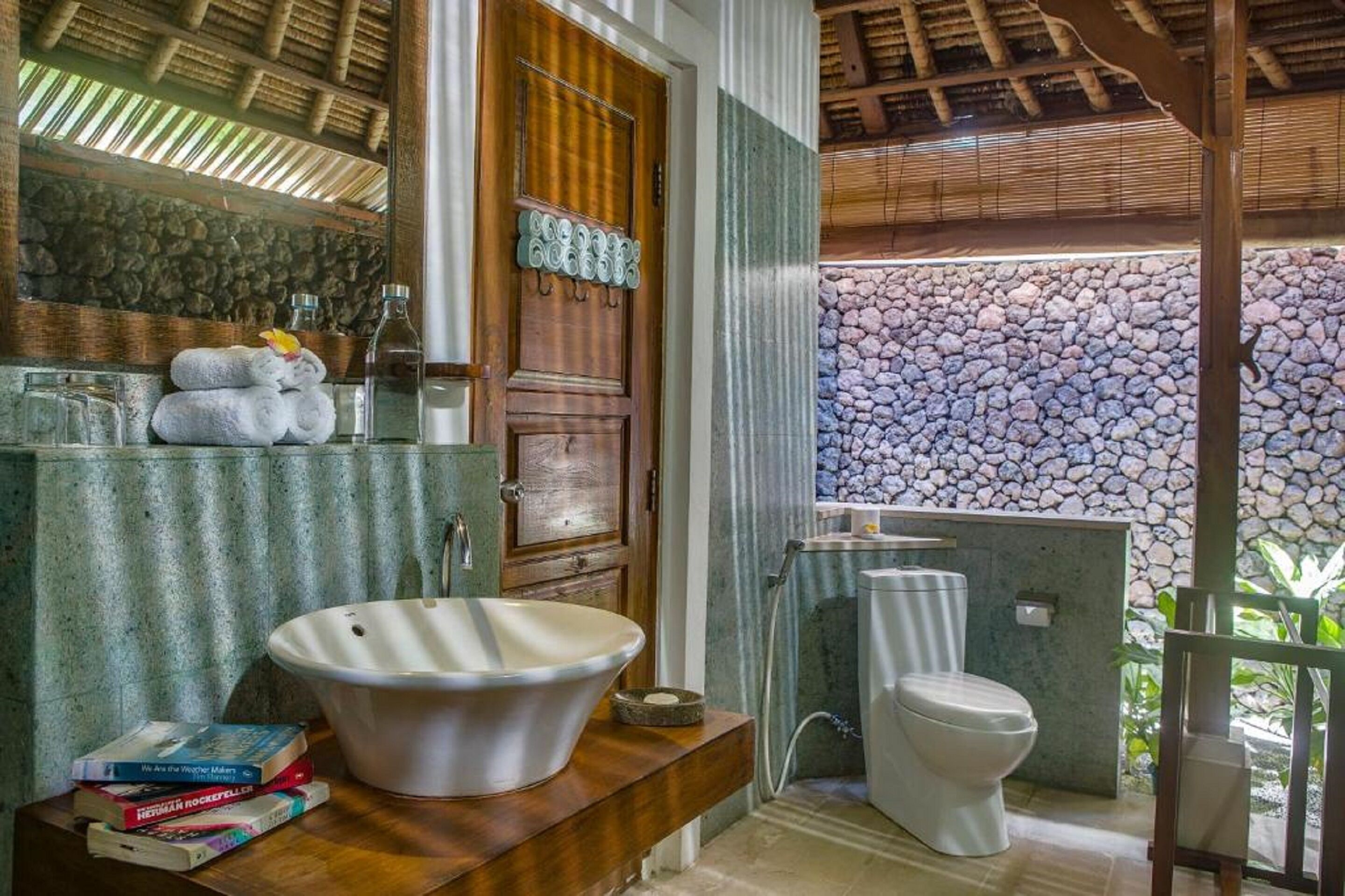 A bathroom with a large white sink, a toilet, and a mirror above the sink. There are towels stacked on the counter and a few books.