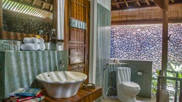 A bathroom with a large white sink, a toilet, and a mirror above the sink. There are towels stacked on the counter and a few books.