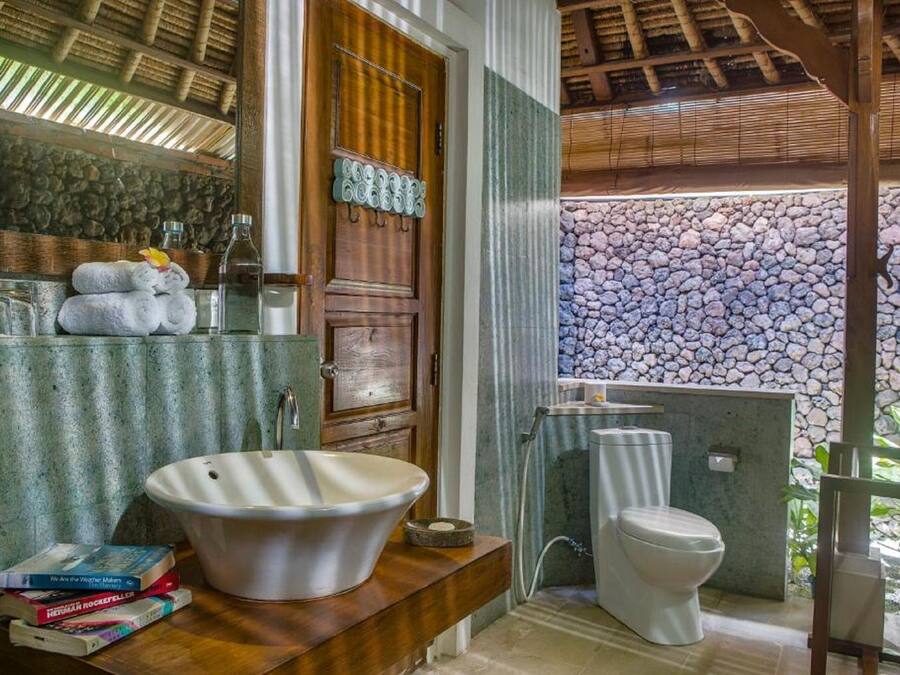 A bathroom with a large white sink, a toilet, and a mirror above the sink. There are towels stacked on the counter and a few books.