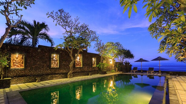 A poolside area with a stone wall, palm trees, and lounge chairs. - Alam Candi Resort (Karangasem)