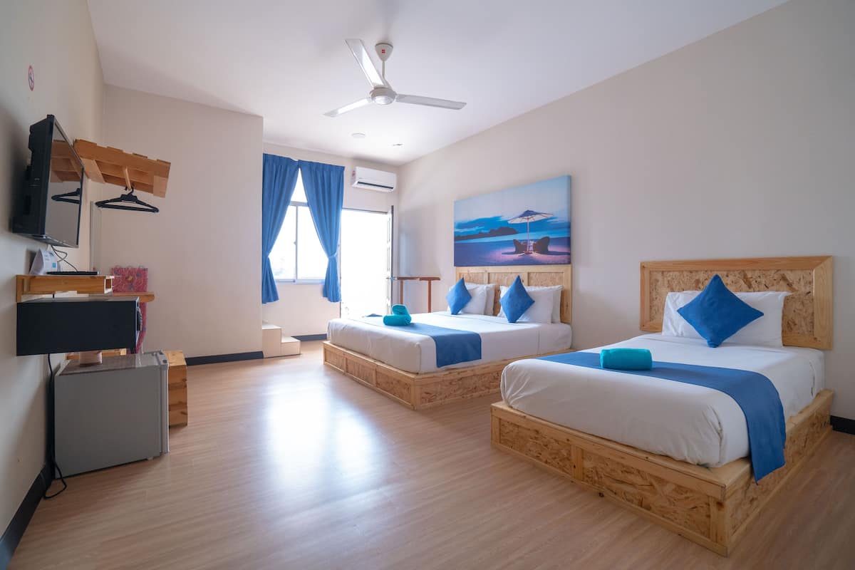 Una camera d'albergo con due letti, una ventola a soffitto, una TV a schermo piatto e un quadro raffigurante una spiaggia.