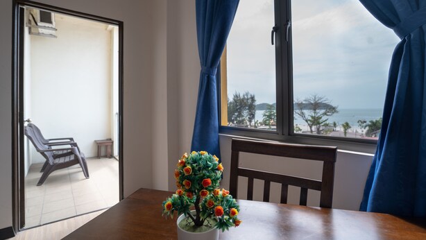 Una habitación con una mesa de madera, una planta en maceta, una silla y vista al mar a través de una ventana con cortinas azules.