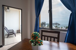 A room with a wooden table, a potted plant, a chair, and a view of the sea through a window with blue curtains. - Paretto Seaview Hotel (Langkawi)