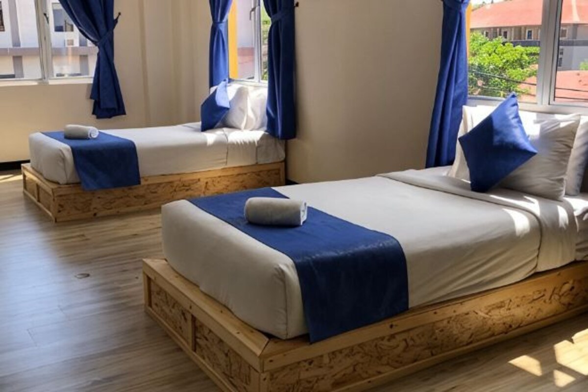 Two wooden beds with blue bedding, a window with blue curtains, and a view of buildings outside.