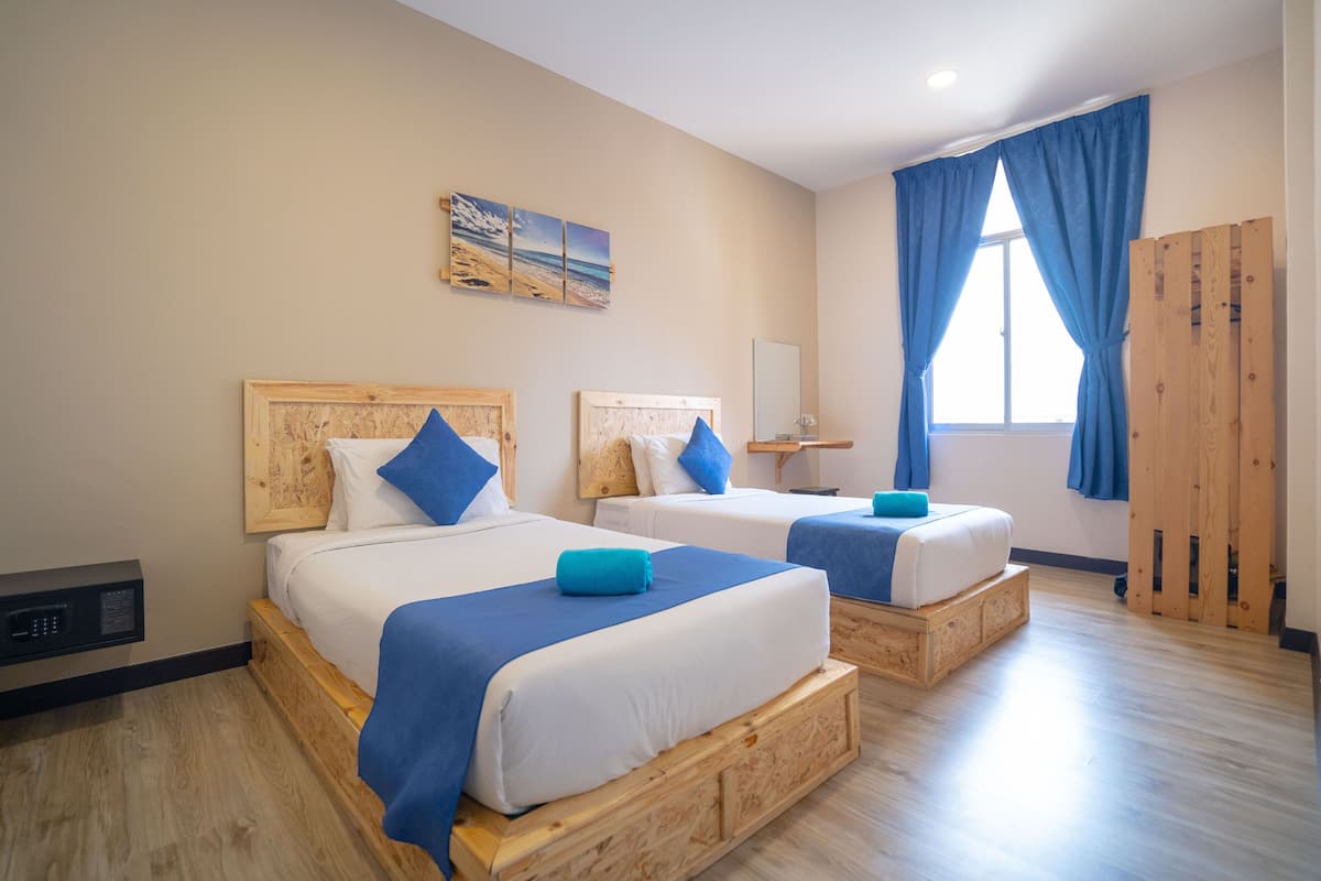 Camera d'albergo con due letti, testiere in legno, un'ampia finestra con tende blu e un quadro a tema spiaggia.