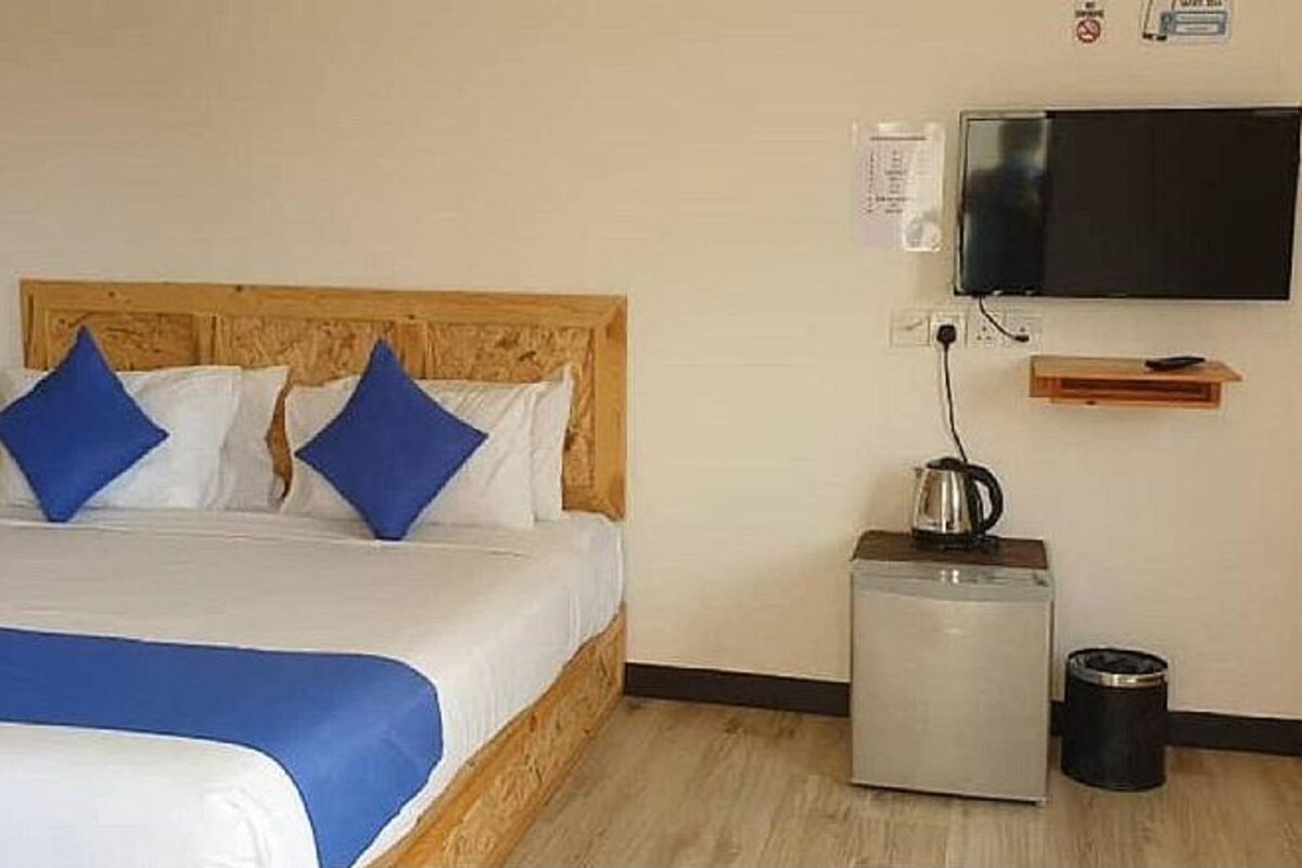 A hotel room with a wooden headboard bed, white and blue bedding, a flat-screen TV mounted on the wall, a small table with a kettle, and a trash bin.