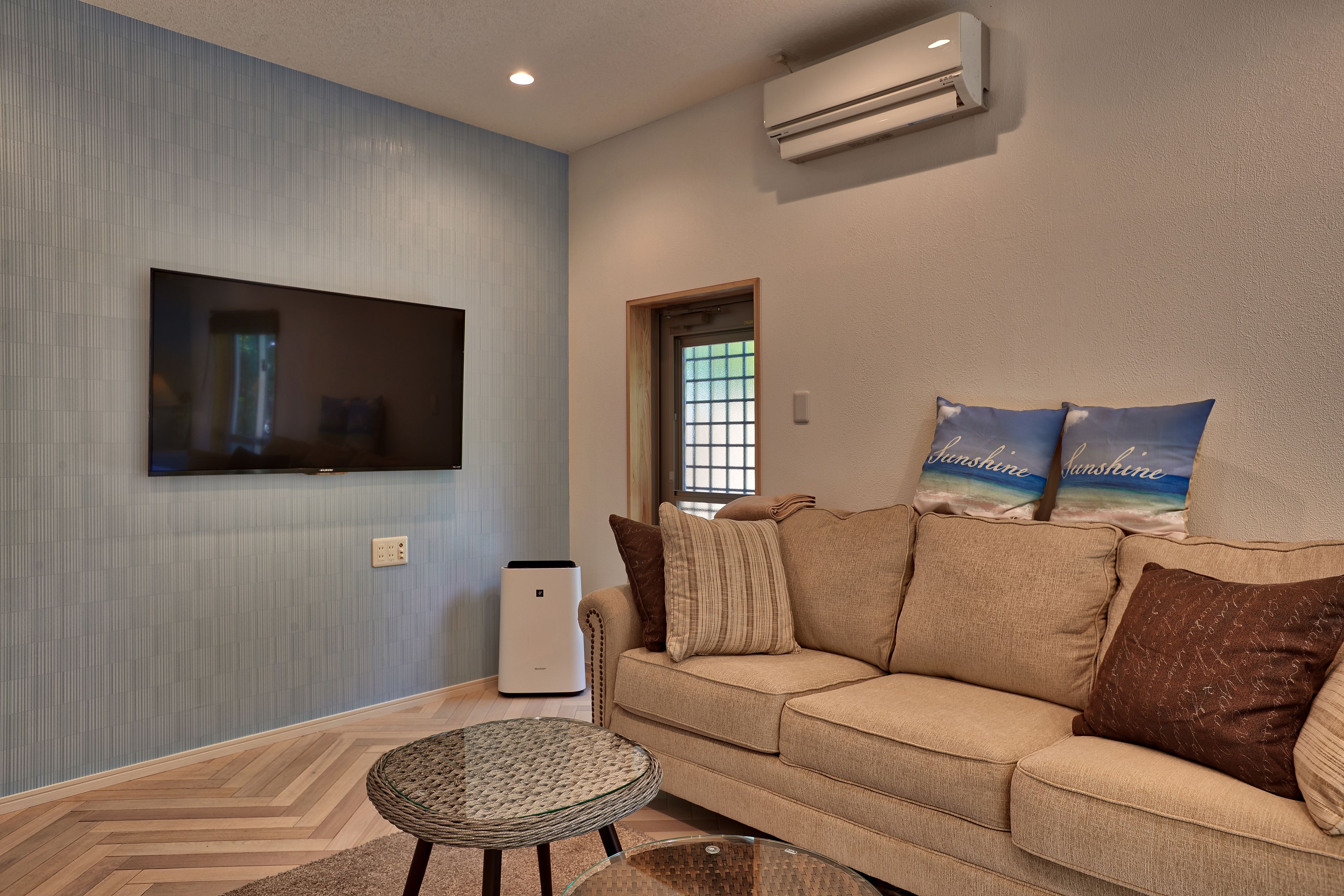 A living room with a beige sofa, a flat-screen TV mounted on the wall, and a small round table.