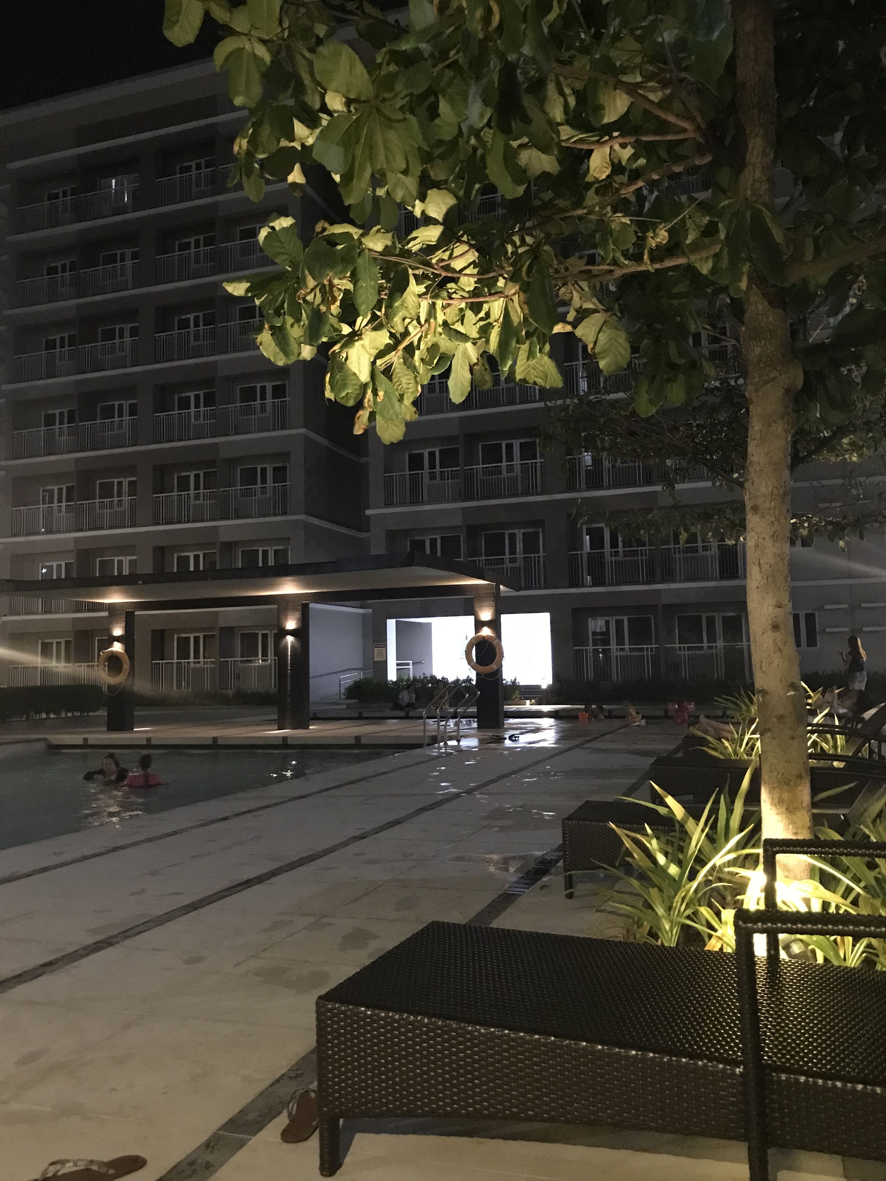 A nighttime outdoor area with a swimming pool, lounge chairs, and a building with multiple balconies in the background.